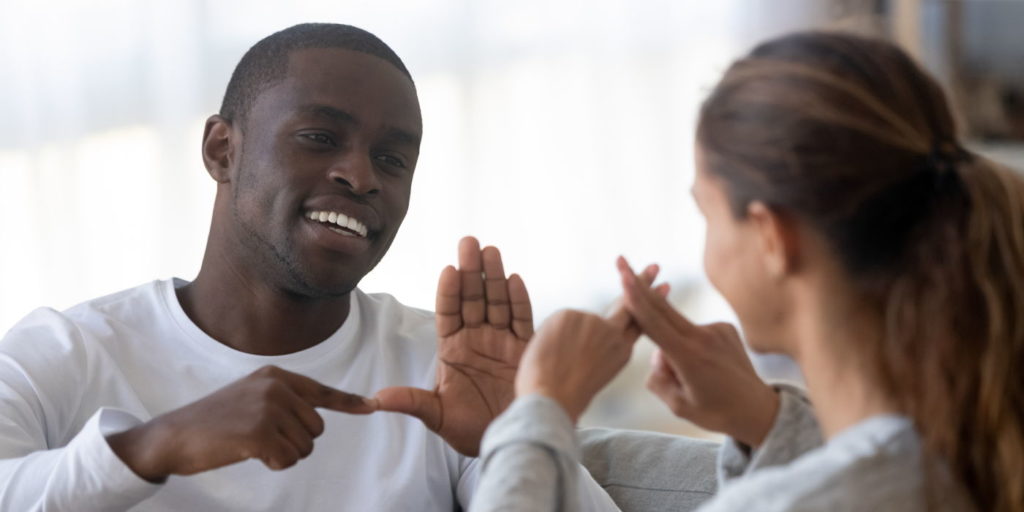 Smiling african American man sit on couch show hand gestures talking with female friend at home, international disabled hearing impaired couple or spouses use sign language communicating